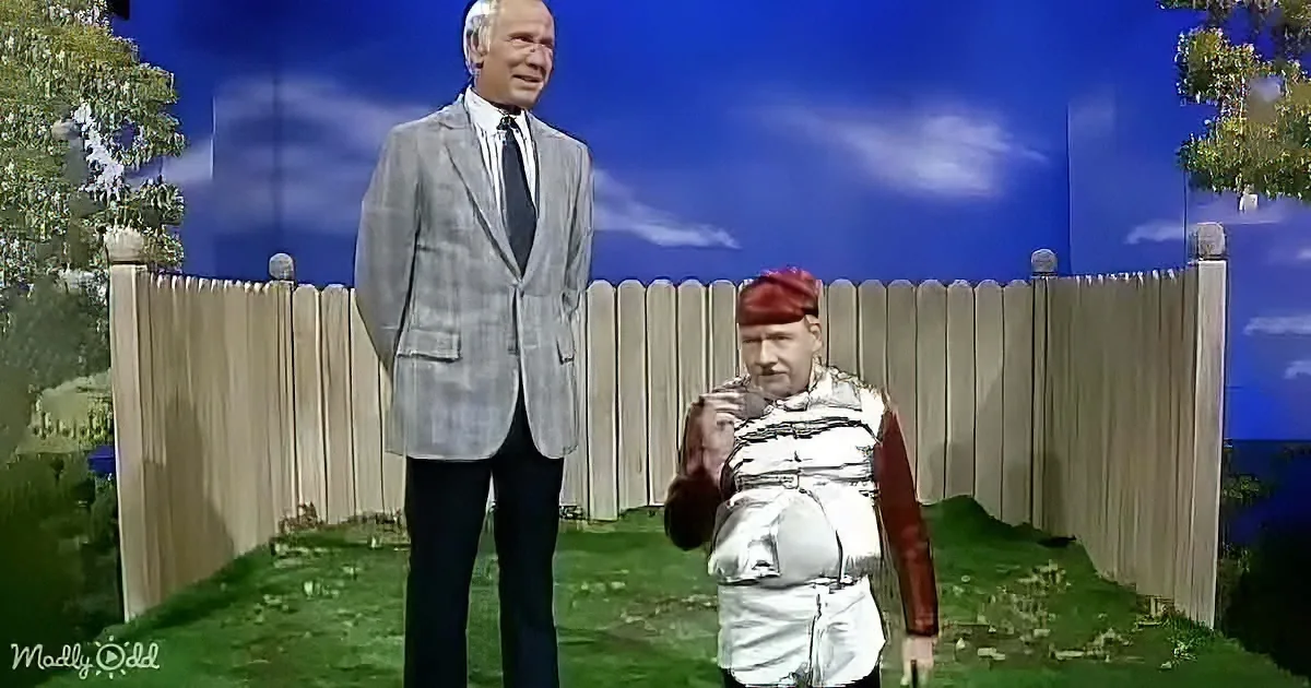 A man in a suit stands next to a person in a silver outfit, both in a backyard setting with a fence and trees.