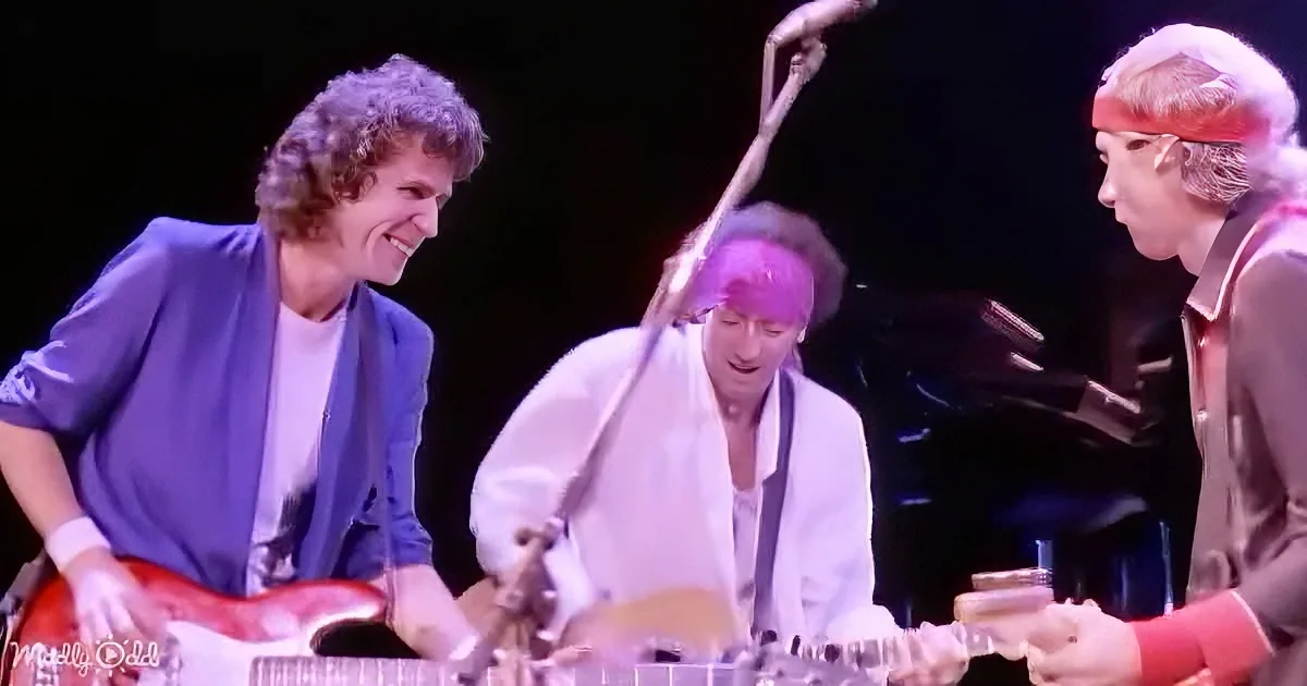 A musician in a blue shirt smiles while playing a red guitar on stage.