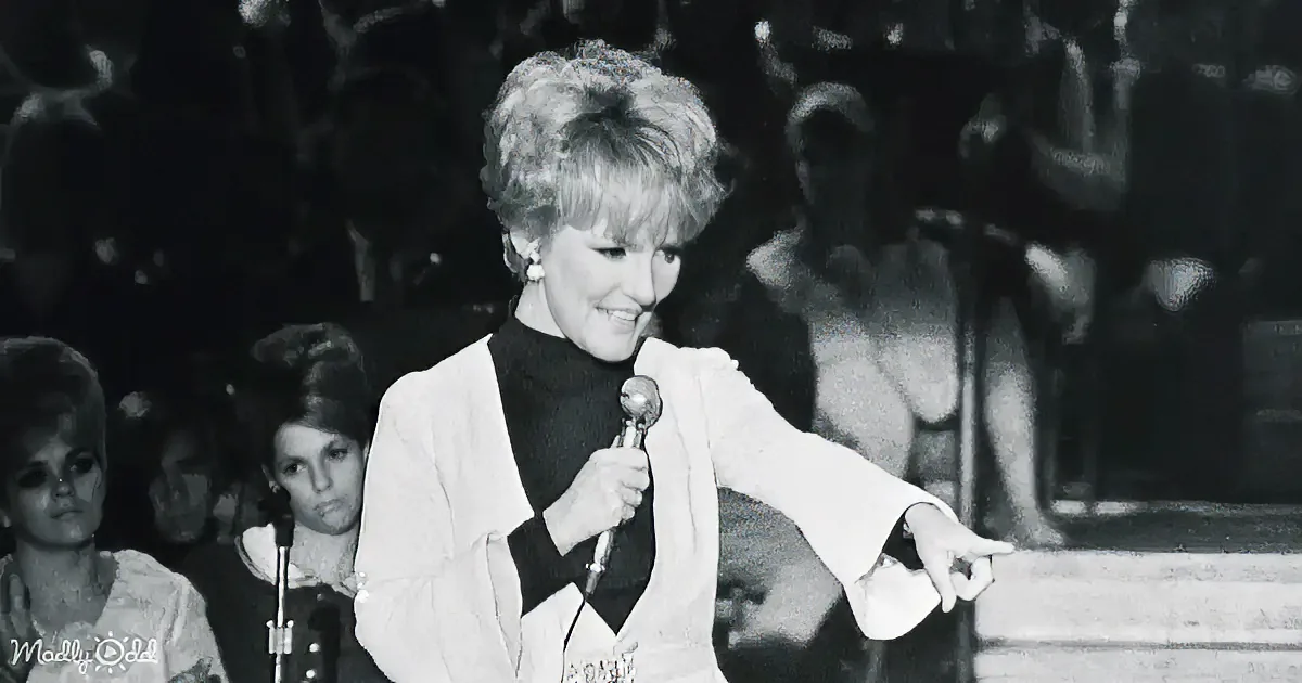 A woman in a white jacket and black turtleneck holds a microphone, smiling, with a band and audience in the background.