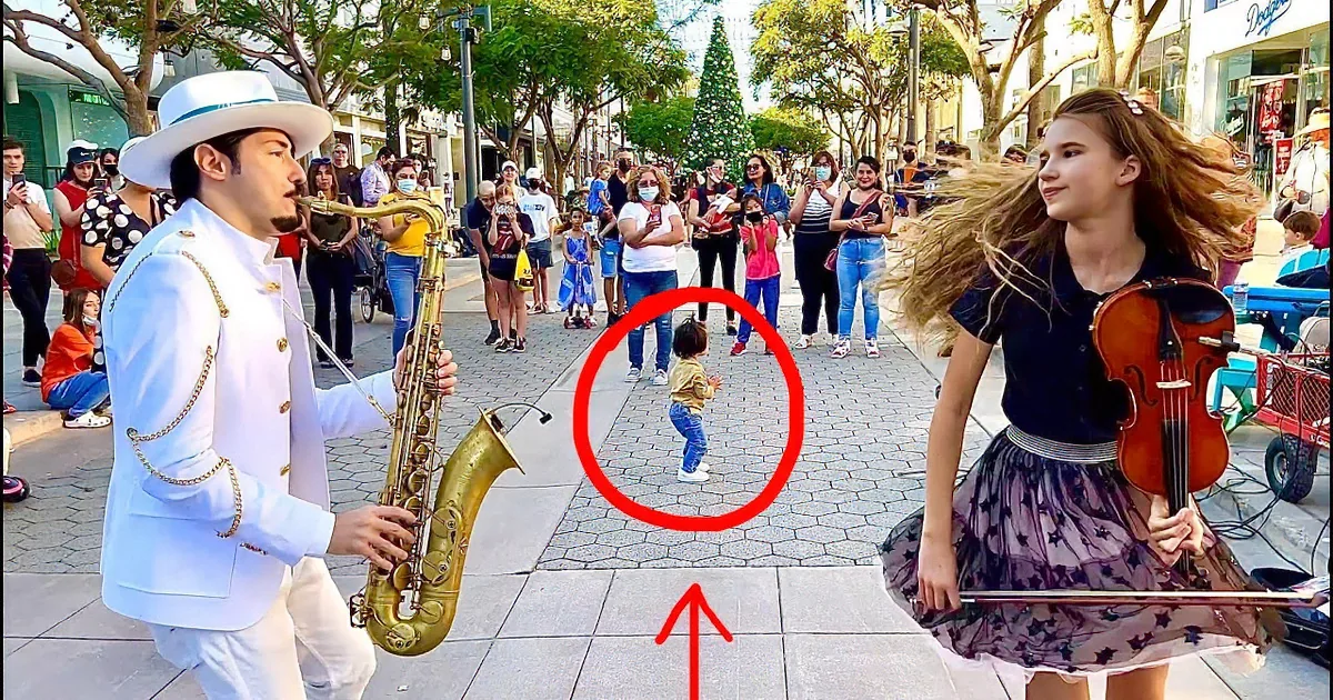 A man in a white suit plays saxophone while a woman in a black dress plays violin on a city sidewalk.