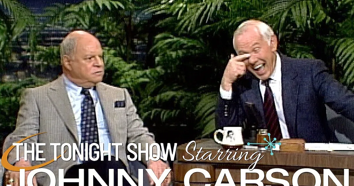 Two men in suits, one laughing, the other looking serious, in a studio setting with plants in the background.