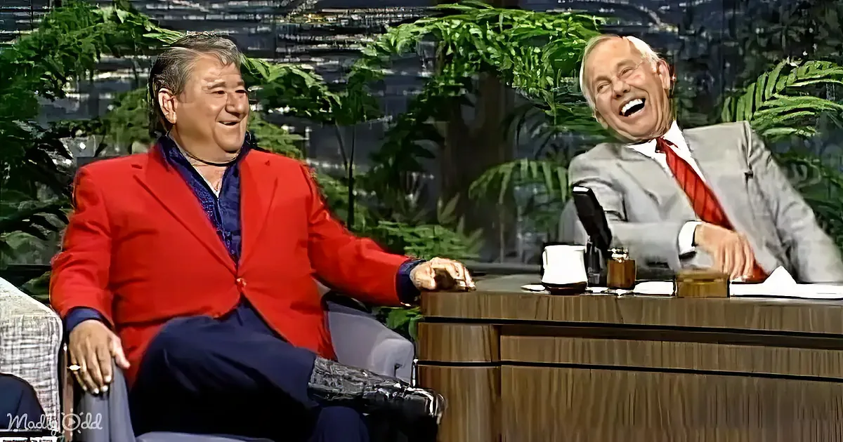 Two men, one in a red suit, are seated and laughing on a talk show set with a backdrop of greenery.