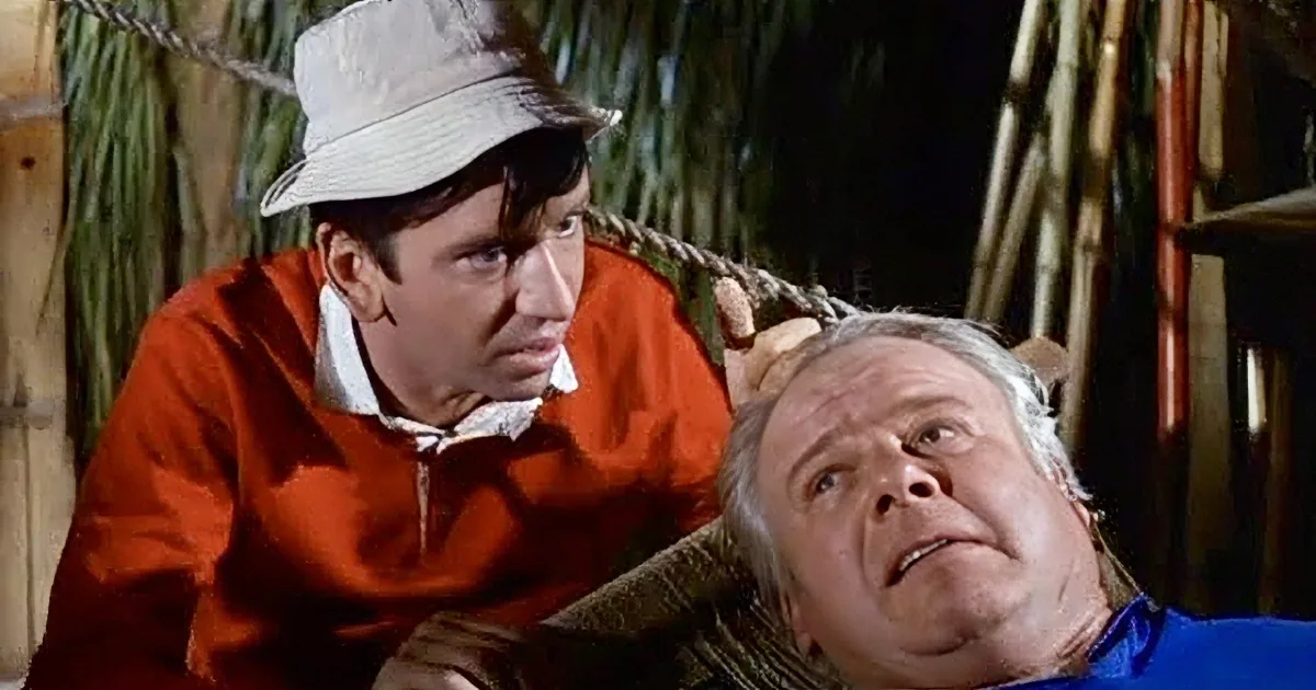A man in a red jacket and hat leans over another man in a blue shirt, both in a rustic setting.