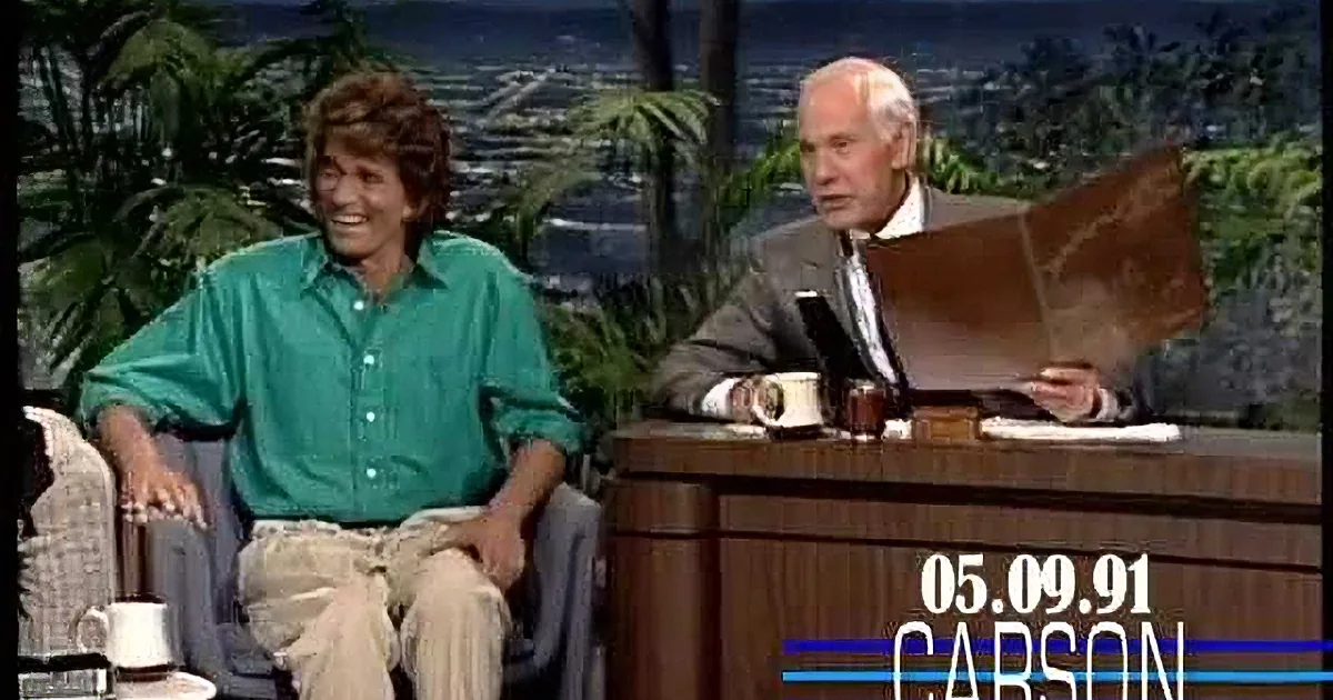 A man in a green shirt smiles while sitting next to a man in a suit at a desk with a mug.