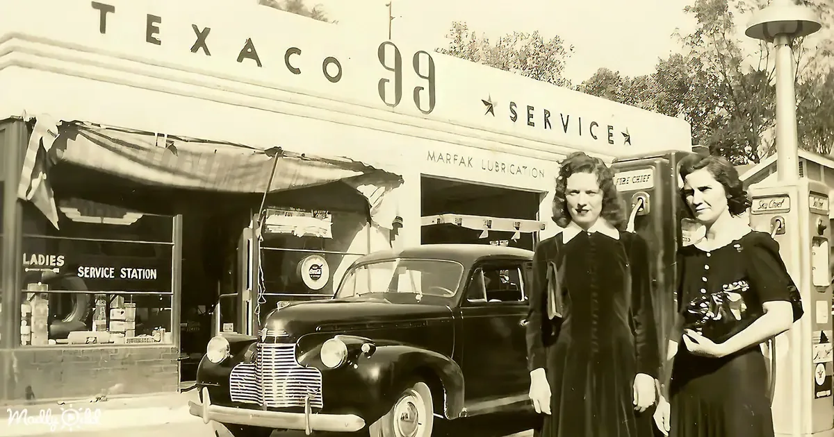 Two women stand beside a vintage car in front of a Texaco 99 Service Station.