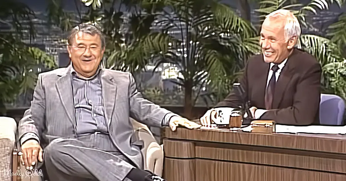 Two men in suits are seated, smiling, with a wooden desk and tropical plants in the background.