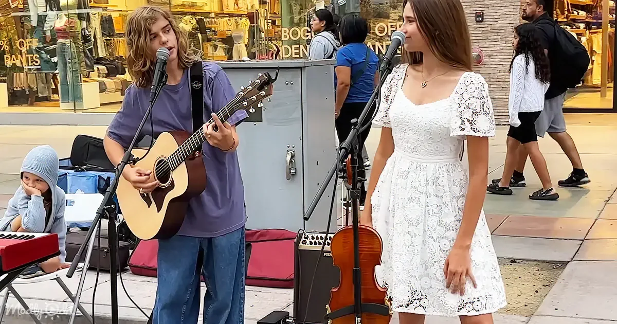 A young man plays guitar while a young woman sings in a white dress, both on a street performance setup.