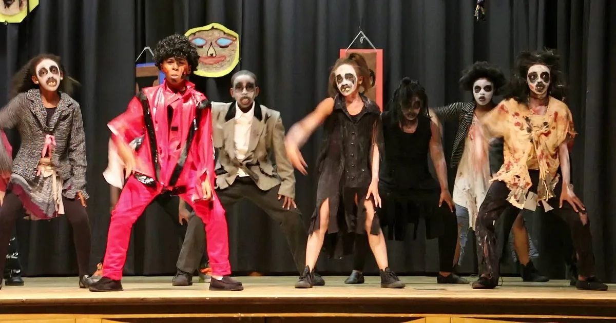 A group of performers in zombie costumes dance on stage, with one in a red outfit leading the group.