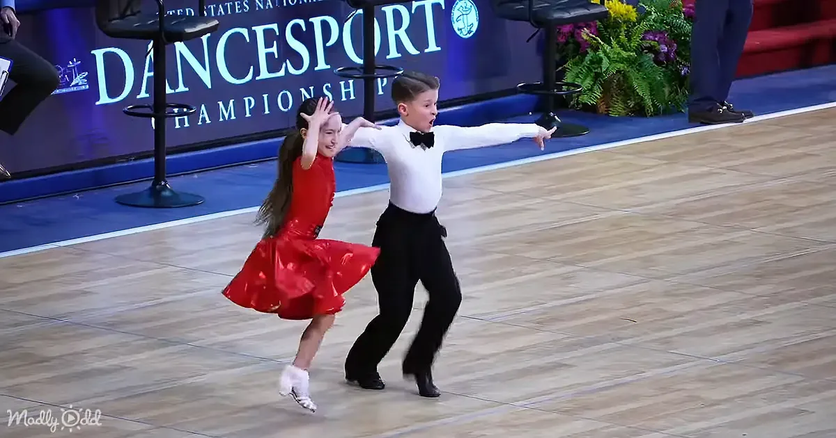 A young boy and girl in formal attire dance gracefully on a dance floor at a competition.