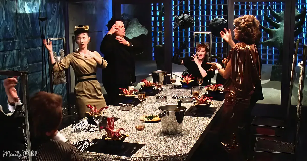 A group of people in a dimly lit room with a table full of food and drinks, one person in a gold outfit appears to be...
