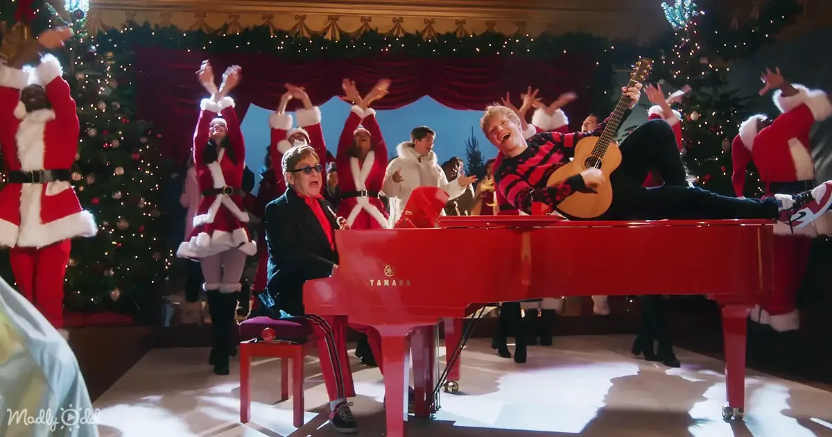 A person in a red and black striped shirt plays guitar on a red piano, surrounded by people in Santa outfits.
