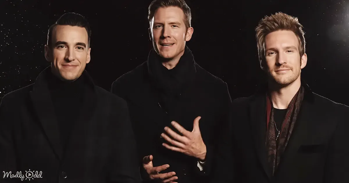 Three men in black attire stand together, smiling and gesturing, against a dark background.
