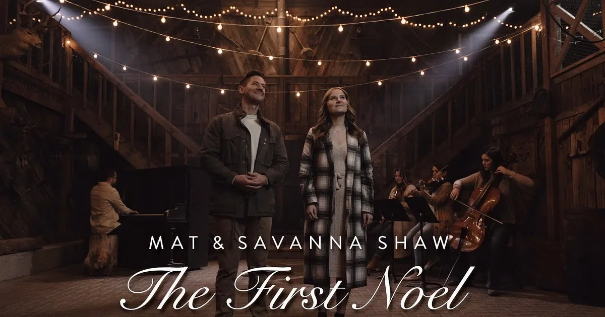 A man and woman stand in a rustic barn with string lights, surrounded by musicians playing instruments.