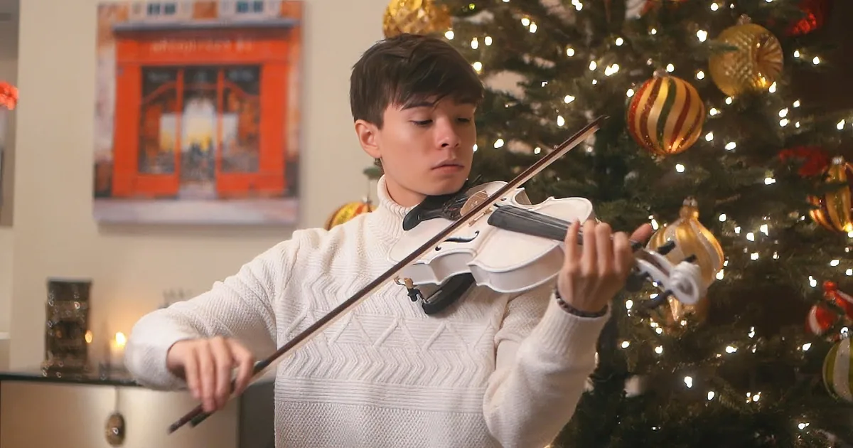 A person in a white sweater plays the violin in front of a decorated Christmas tree.