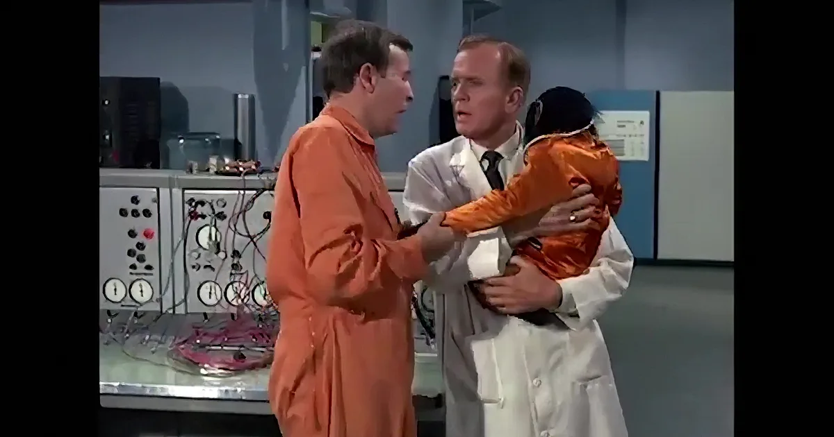 A chimp in an orange jumpsuit is holding a phone to its ear in a room with wires and equipment.