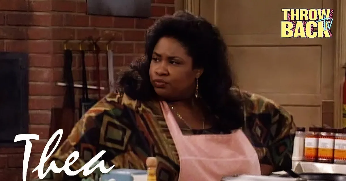 A man in a pink shirt and a woman in a patterned shirt and apron are having a conversation in a kitchen setting.