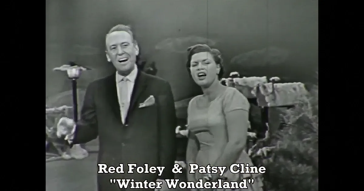A man in a suit and a woman in a dress stand together, possibly on a stage, with a scenic backdrop.