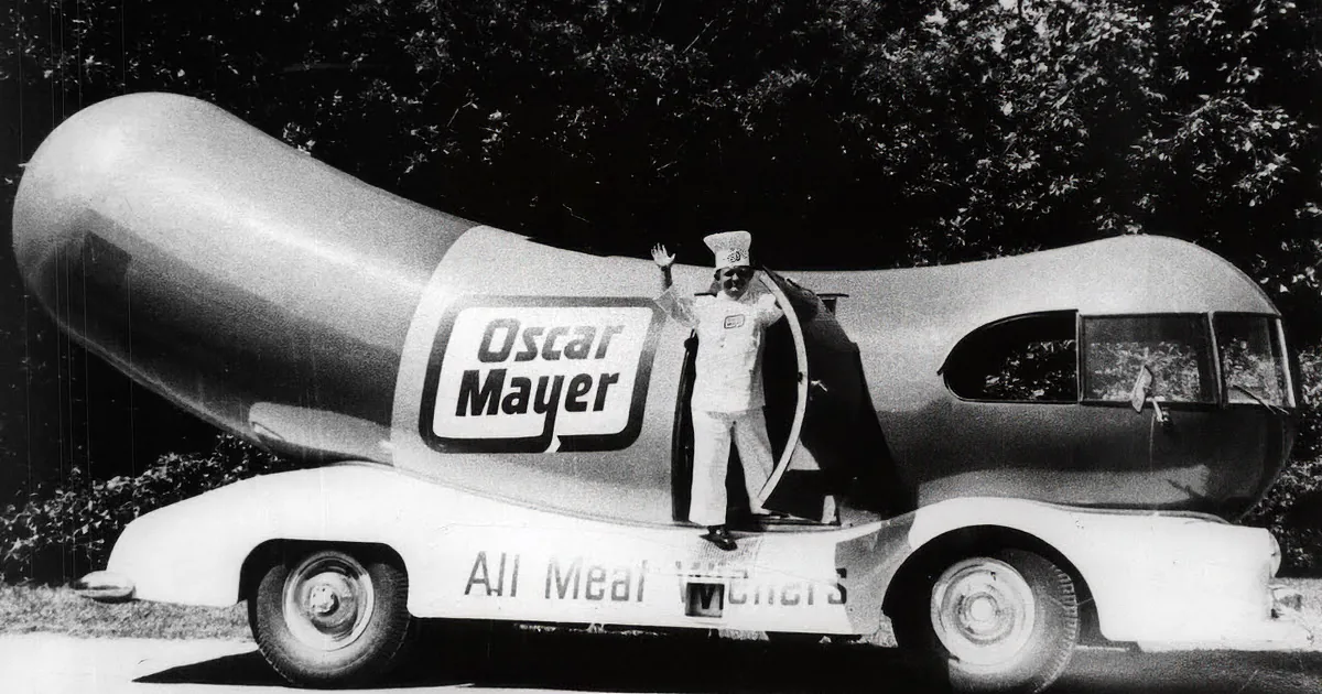 A man stands beside a giant hot dog-shaped vehicle with children nearby, celebrating.