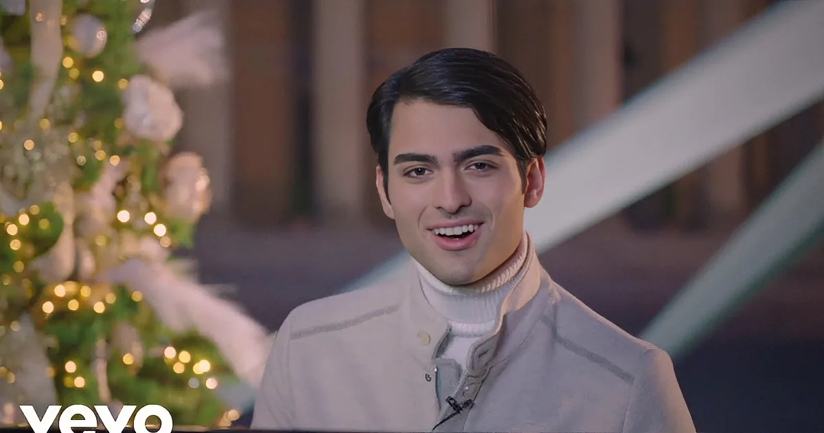 A smiling man in a white turtleneck sweater stands in front of a decorated Christmas tree with lights and ornaments.