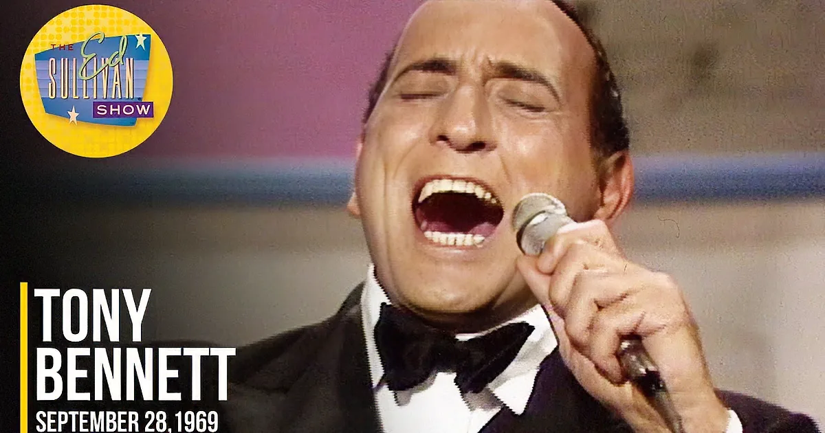 A man in a tuxedo smiles while holding a microphone, wearing a bow tie.