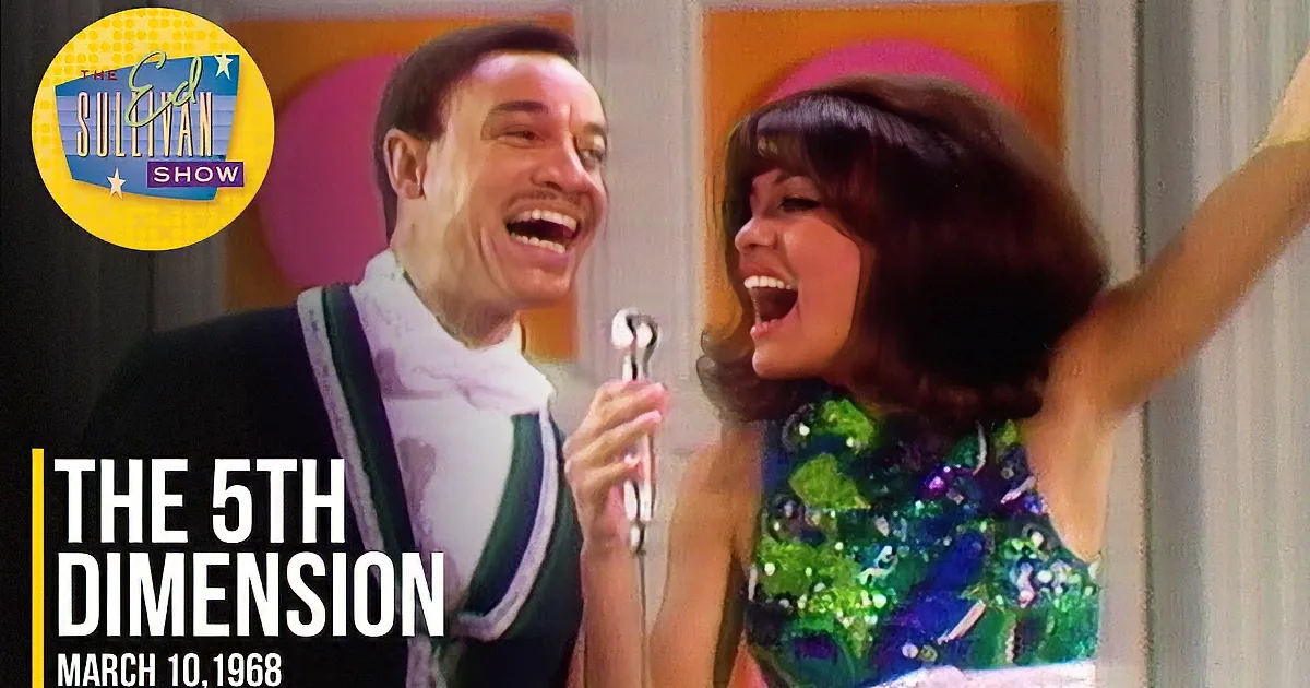 A man and woman are singing together, the woman holding a microphone and wearing a colorful outfit.