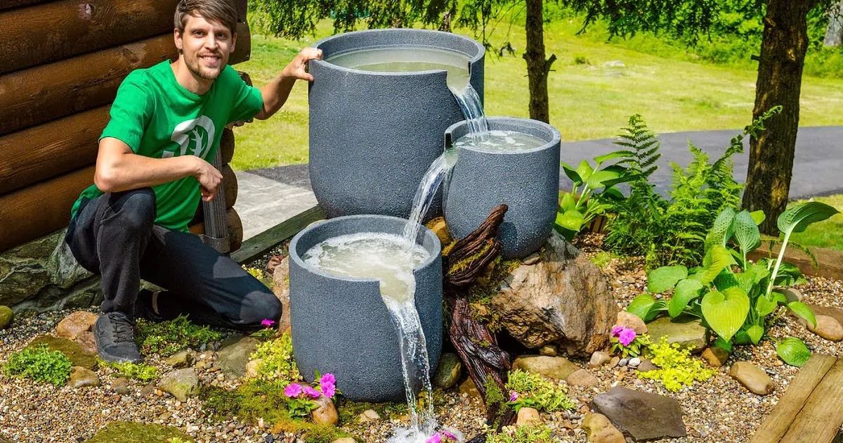 A man in a green shirt kneels beside a cascading water feature in a garden setting.