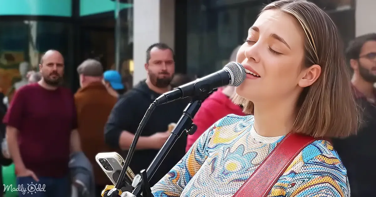 A woman sings passionately into a microphone, wearing a colorful patterned shirt, with a crowd and buildings in the...