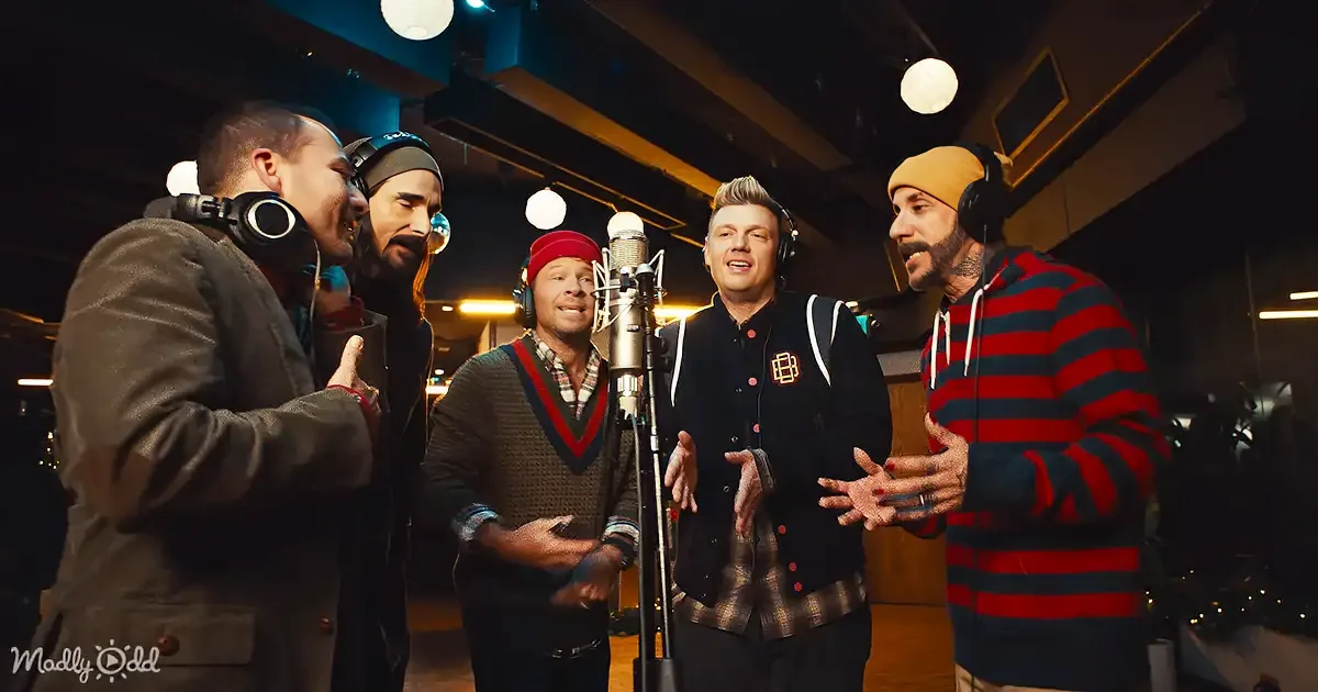 Five men in a recording studio, one singing into a microphone, others listening and smiling.
