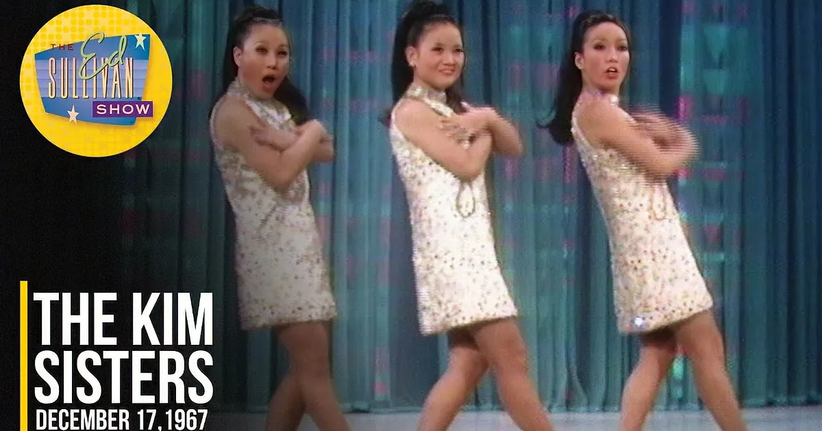 Three women in sequined dresses perform synchronized hand gestures against a blue curtain backdrop.