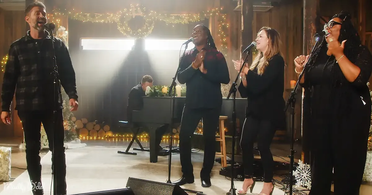 A group of four people sing on stage with microphones, accompanied by a pianist in the background.