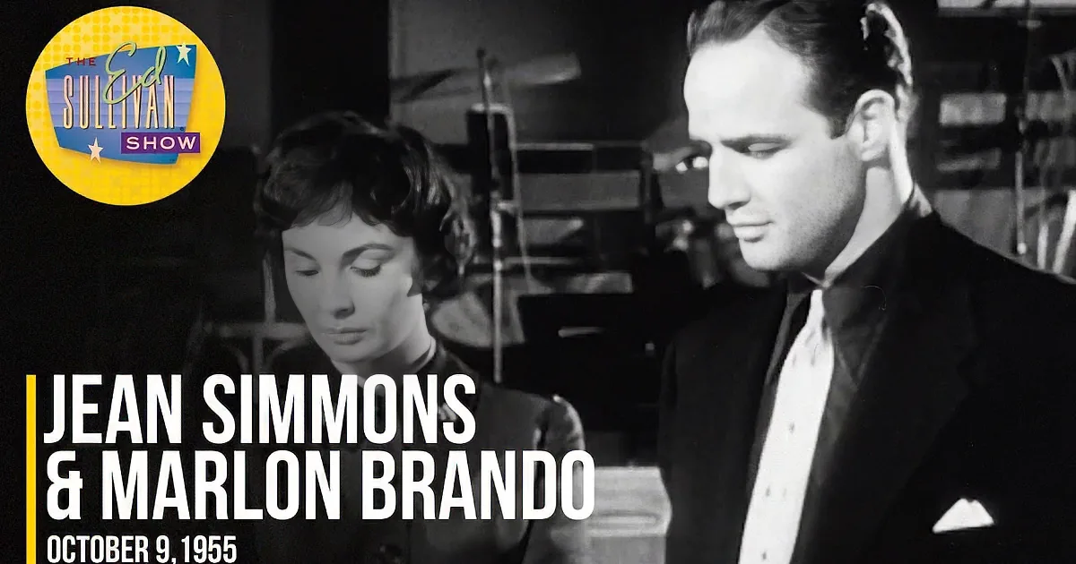 A woman in a black dress with a man in a suit, both looking down, in a dimly lit room with chairs and equipment.