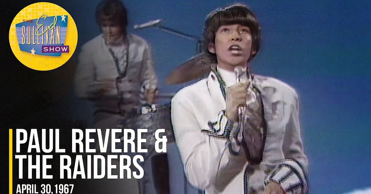 A person in a white outfit sings into a microphone, with another person in the background.