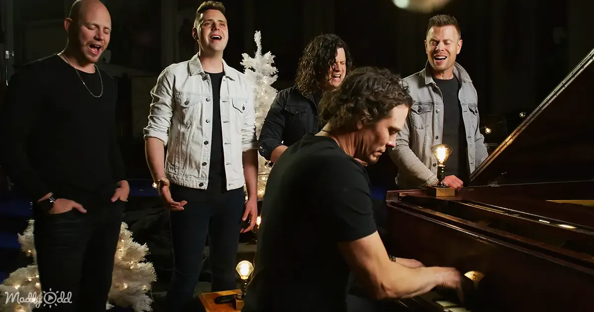 A man plays piano while five others sing and smile around him, with Christmas trees and lights in the background.