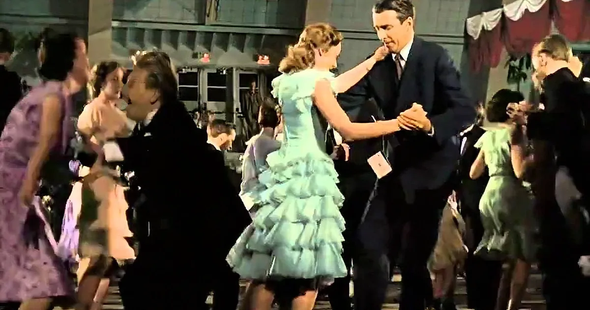 A man in a suit dances with a woman in a light blue dress, surrounded by other dancers in a lively setting.