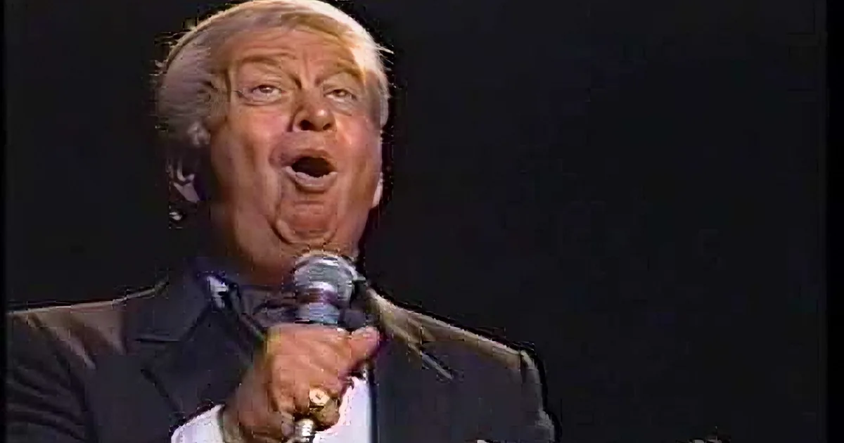 A man in a suit and bow tie speaks into a microphone against a dark background.