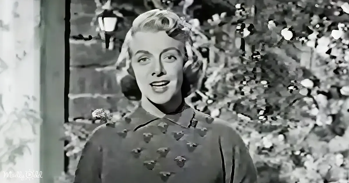 A woman in a floral dress stands amidst greenery, looking to the side with a slight smile.