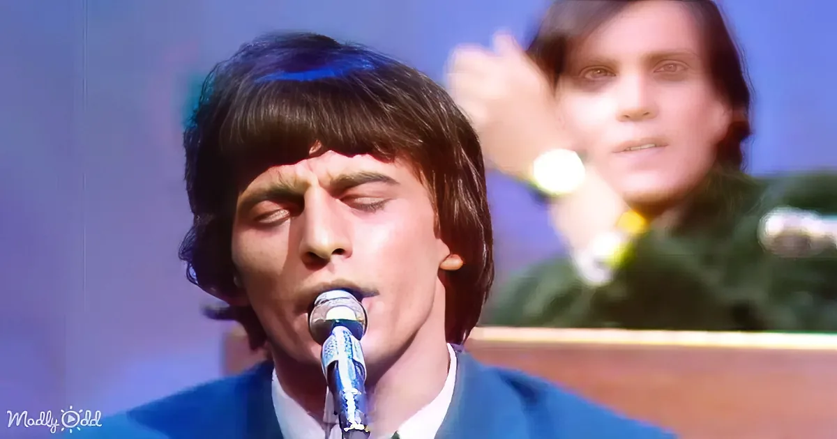A man in a blue suit sings into a microphone with his eyes closed, while another person is blurred in the background.