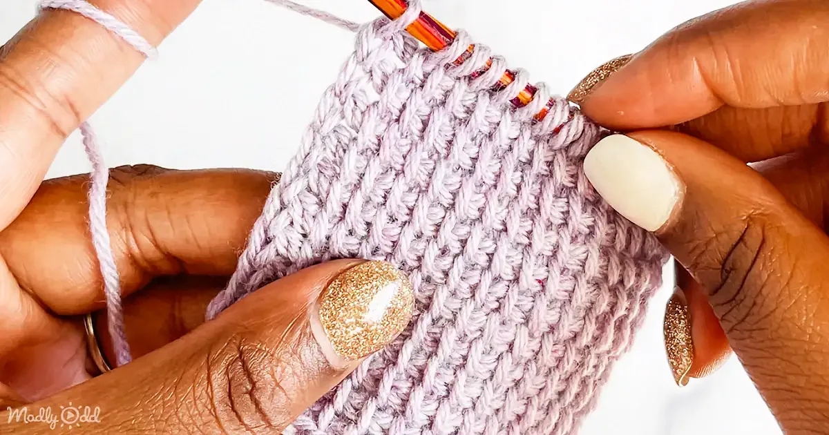 A hand with glittery nails holds a crochet project in progress, showcasing a textured fabric.