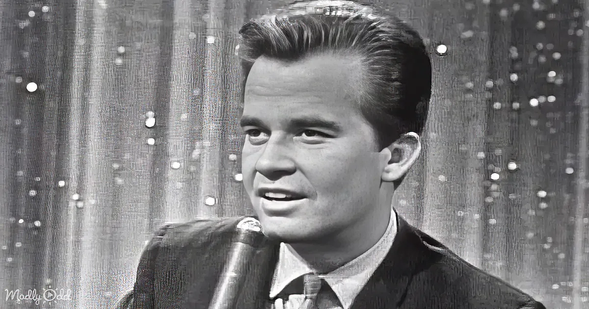A man in a suit speaks into a microphone against a textured backdrop.
