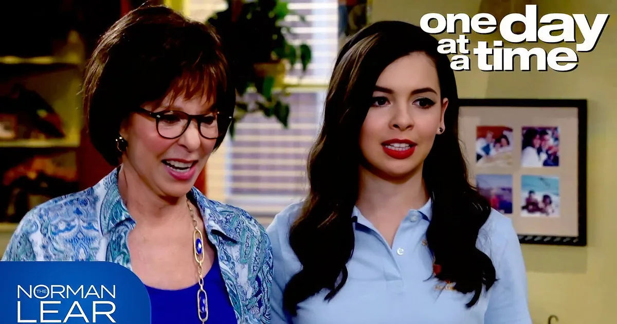 Two women, one in glasses and blue top, the other in light blue shirt, are smiling and standing indoors.