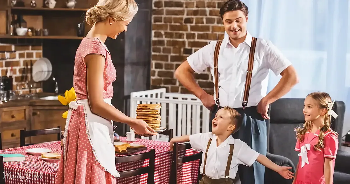 A woman in a polka-dot dress serves pancakes to a boy while a man and girl watch in a cozy kitchen.