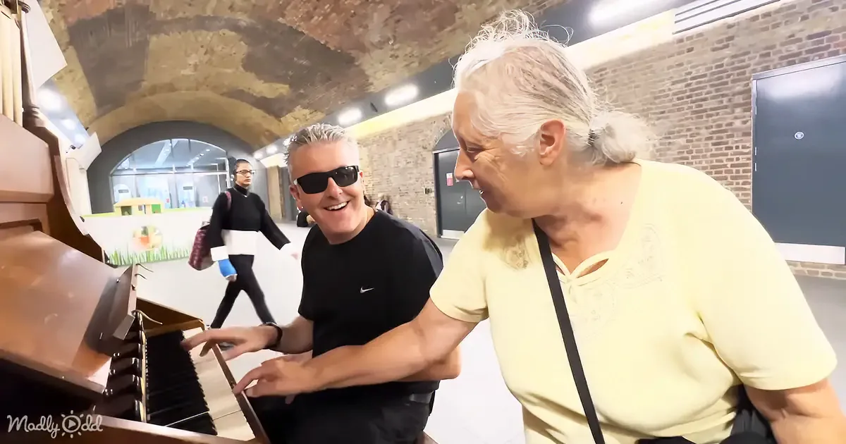 A man in sunglasses plays piano while an older woman leans in, smiling in a brick-walled room.