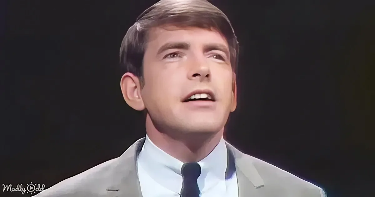 A man in a suit and tie is singing or speaking, looking upwards with a serious expression.