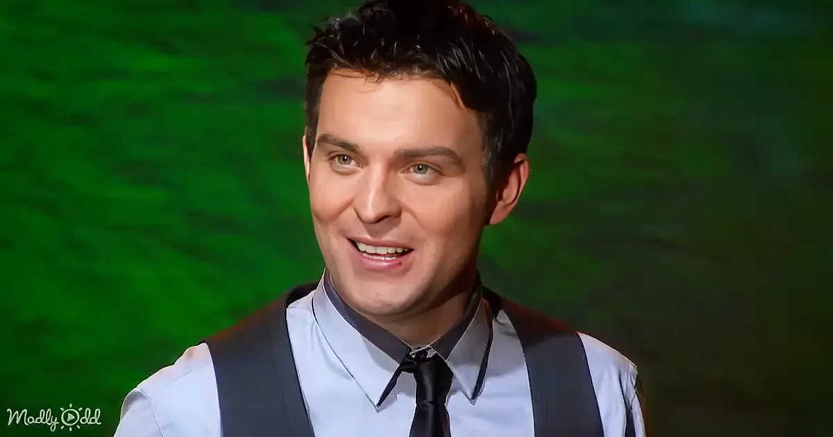 A man in a light blue shirt, black tie, and suspenders smiles against a green background.