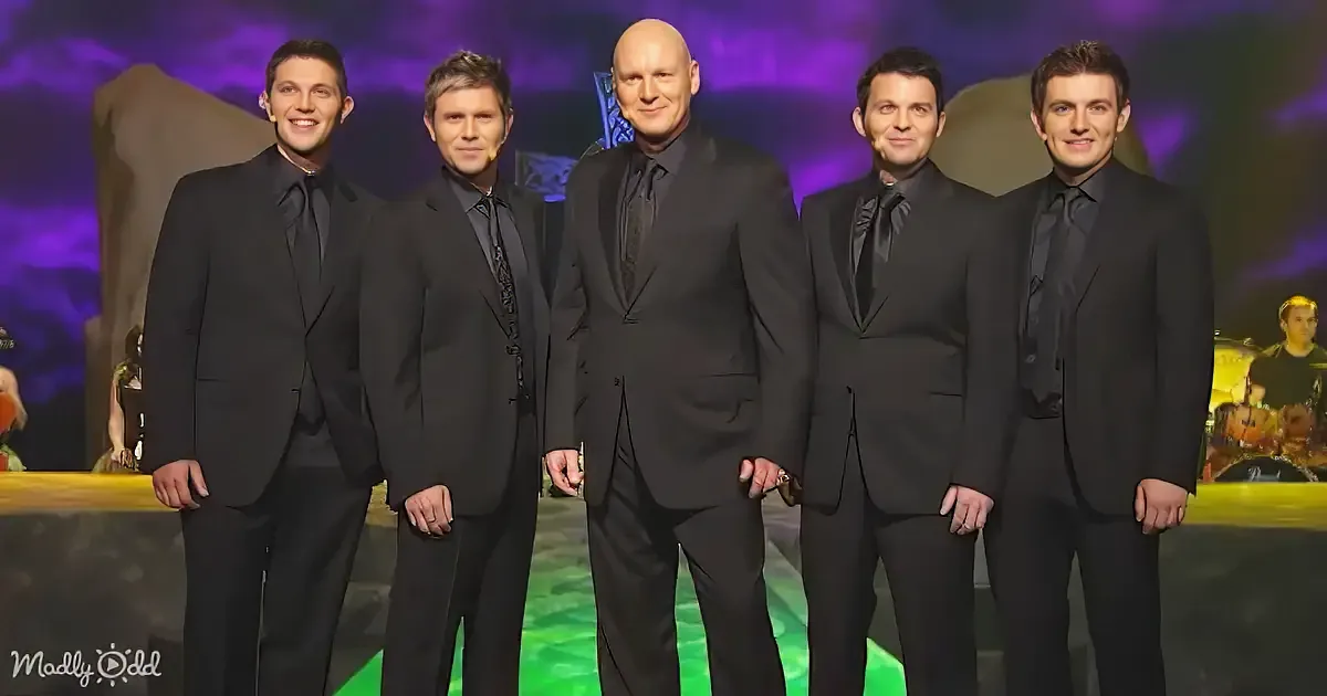 A group of five men in black suits stands together on a stage with a purple and green backdrop.