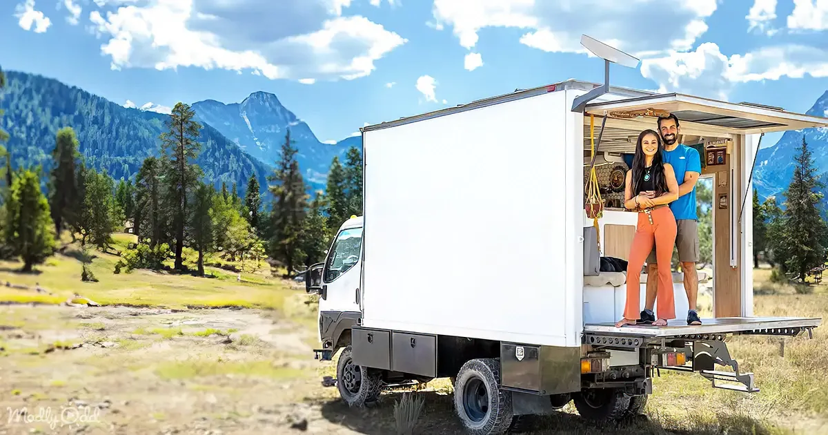 A couple stands on a white camper truck in a scenic mountainous area with trees and a clear sky.