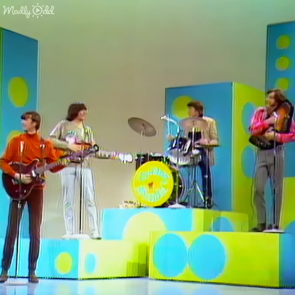 A band performs on a colorful stage with retro, psychedelic decor, including a drum set and guitarists.