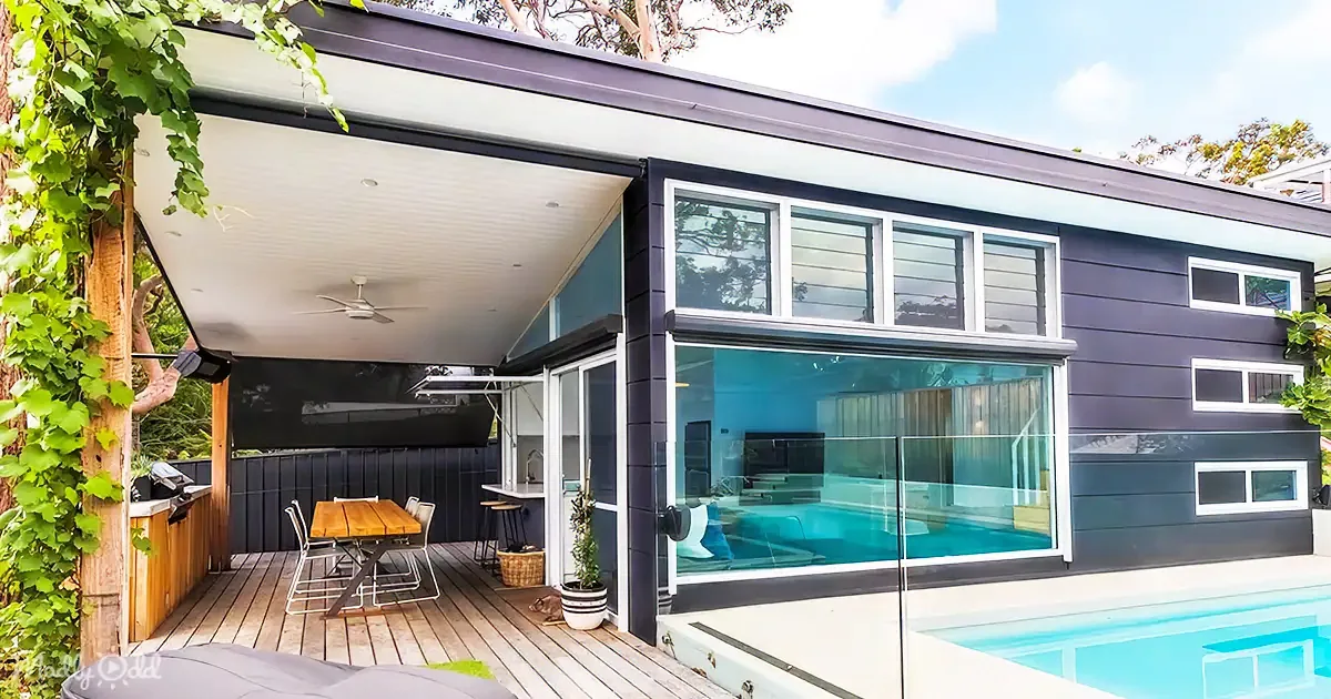 A modern house with a large glass window, a wooden deck, and a pool, surrounded by greenery.
