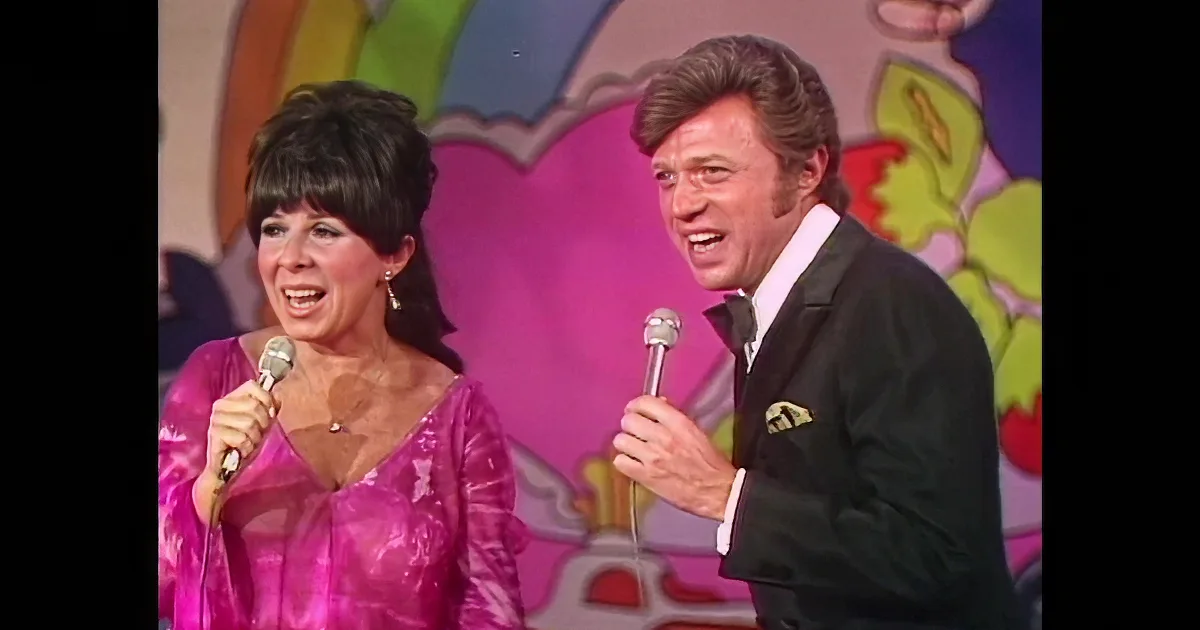 A man and woman in formal attire sing into microphones on a colorful stage backdrop.