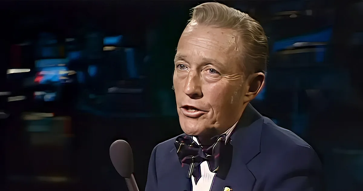 A man in a suit and bow tie speaks into a microphone, with a blurred background suggesting a studio setting.